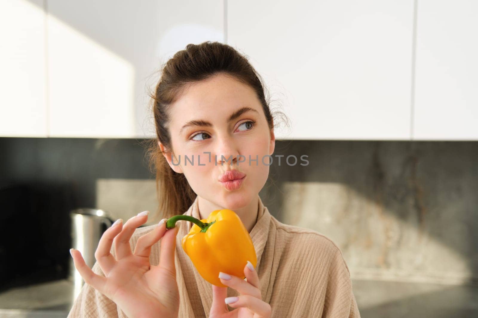 Close up of brunette woman in robe, holding yellow pepper, thinking what to cook, preparing salad or soup, making vegetarian meal with vegetables, standing in kitchen by Benzoix