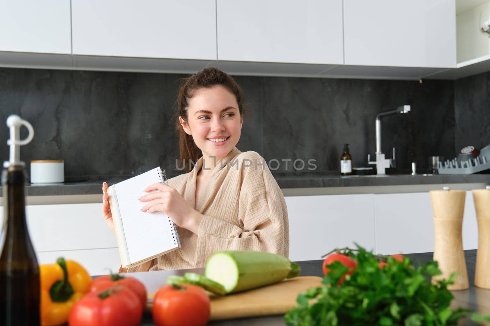 Portrait of woman writing down list of groceries, making notes in recipe, sitting in kitchen near vegetables, preparing dinner menu by Benzoix