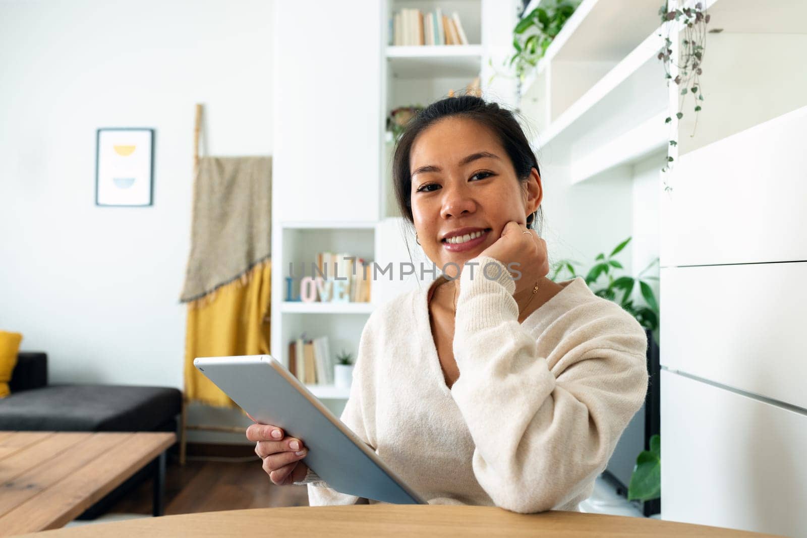 Happy young Asian woman working at home office holding tablet looking at camera. by Hoverstock