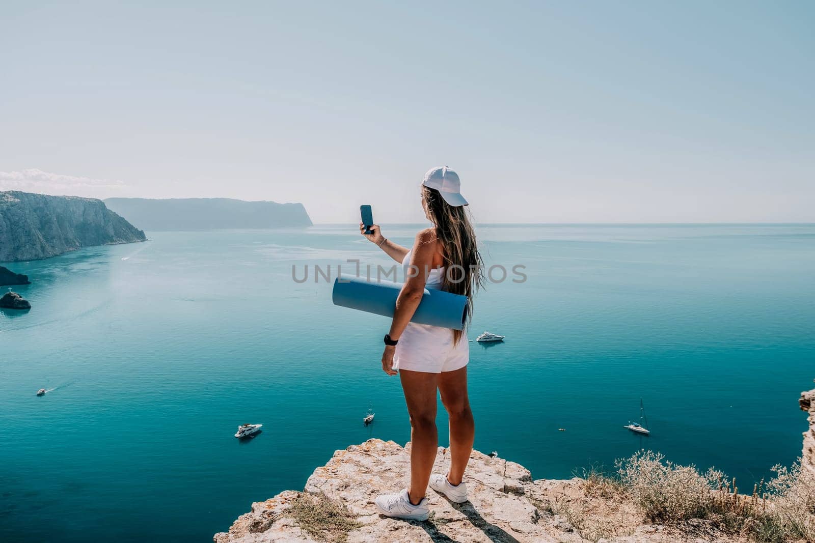 Woman with yoga mat over calm azure sea. Well looking middle aged woman with long hair, fitness instructor with blue yoga mat near the sea. Female fitness yoga routine concept. Healthy lifestyle. by panophotograph