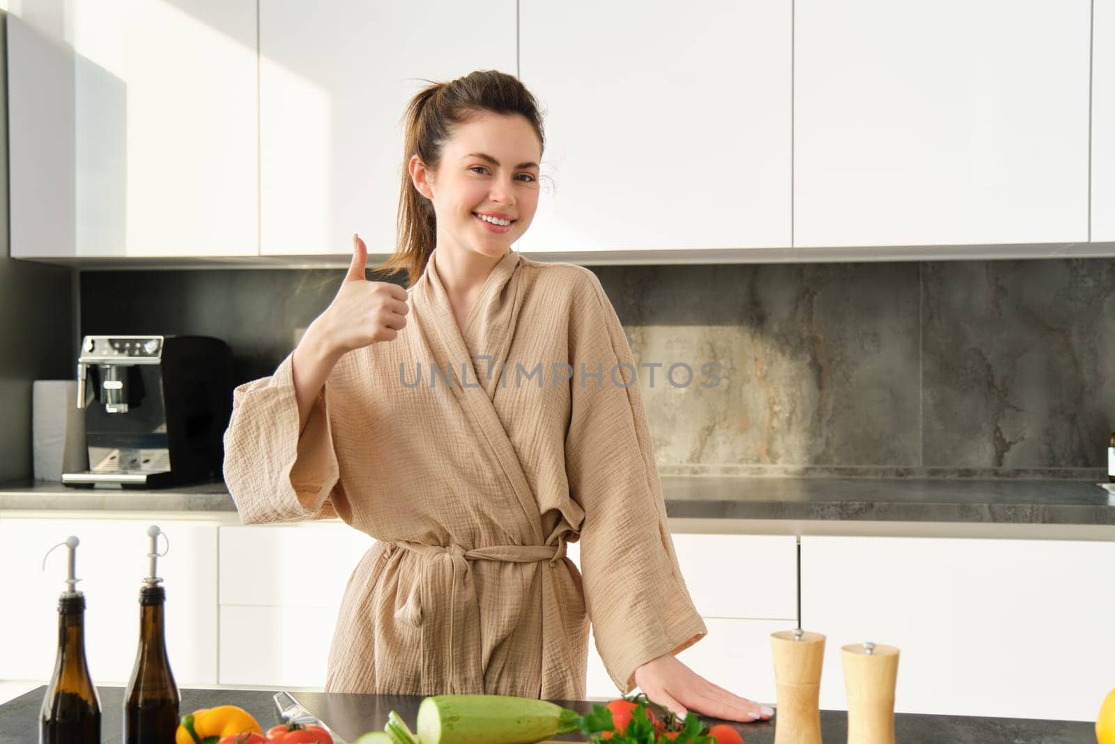 Portrait of smiling woman in bathrobe, shows thumbs up, stands in kitchen, cooking salad, preparing vegetarian dinner, chopping vegetables on board, Food and lifestyle concept