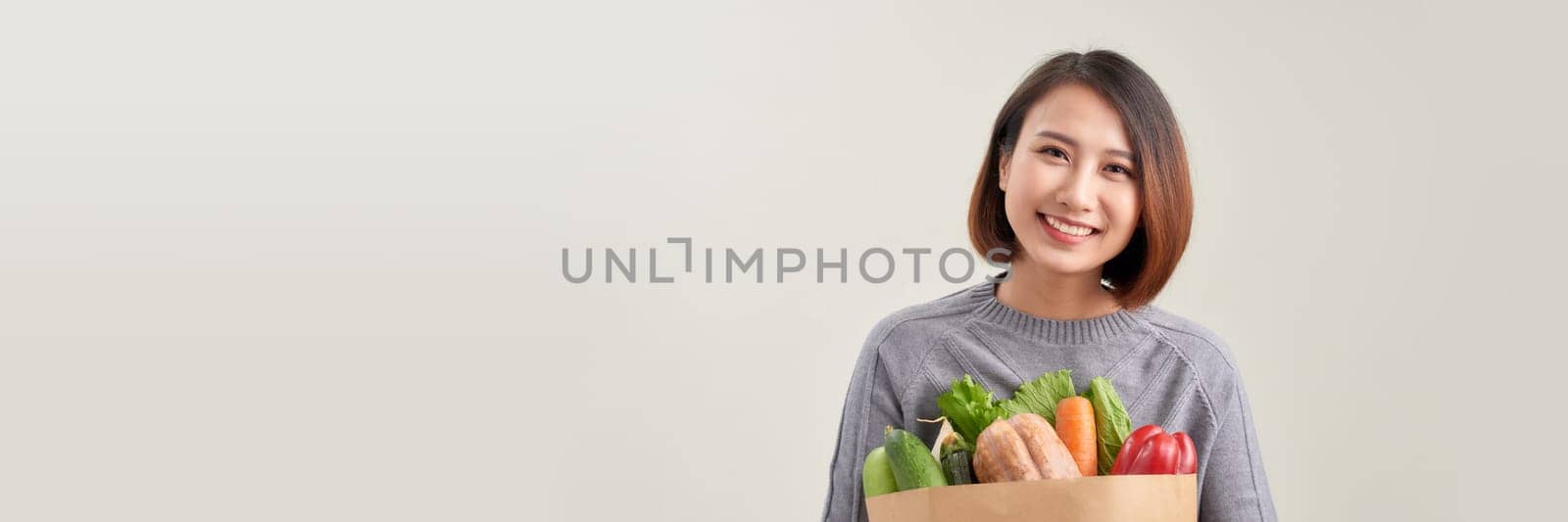 Smiling happy young asian housewife hold paper bag with fresh food products on white background by makidotvn