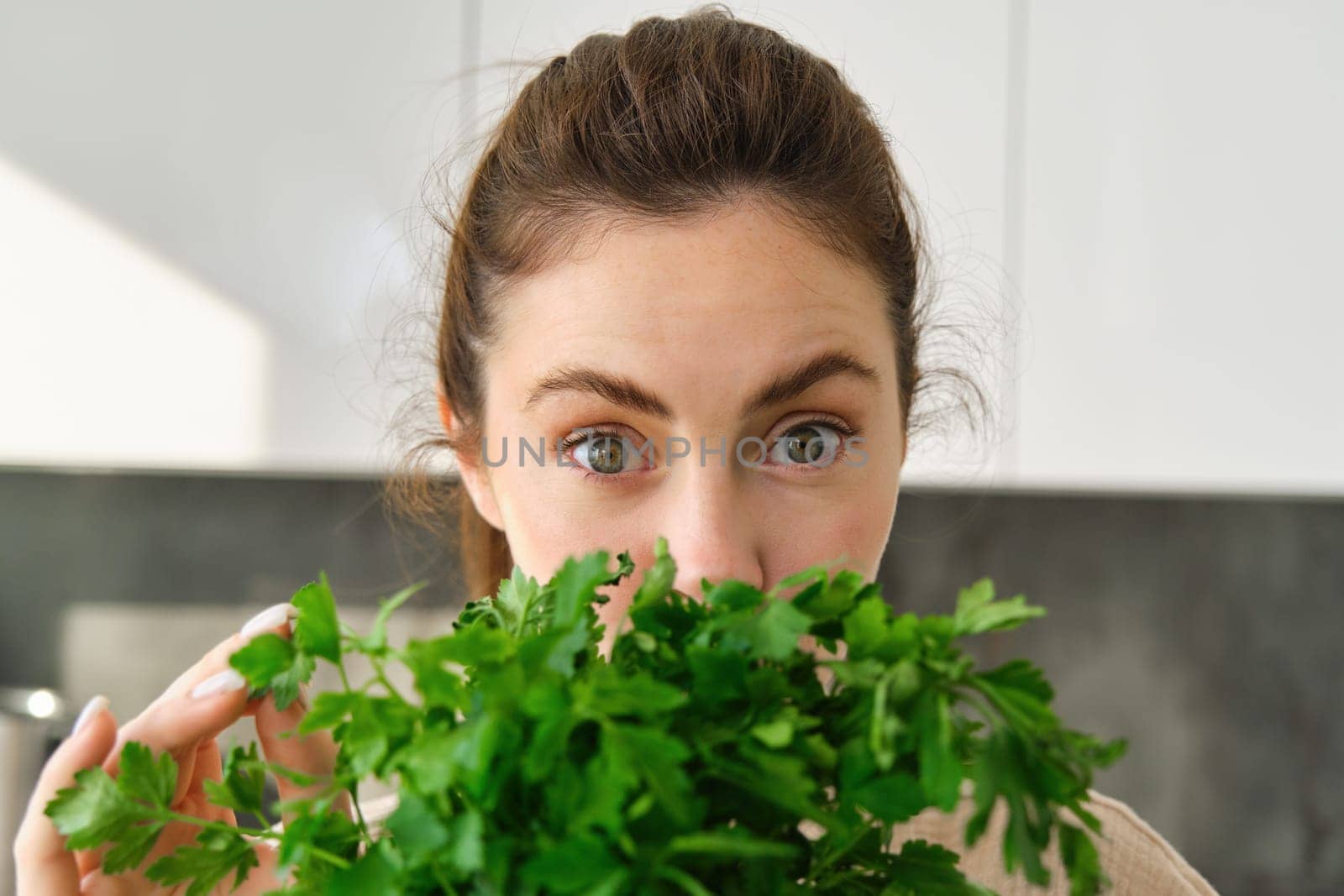Portrait of attractive girl with bunch of parsley, eating fresh herbs and vegetables, cooking in the kitchen.