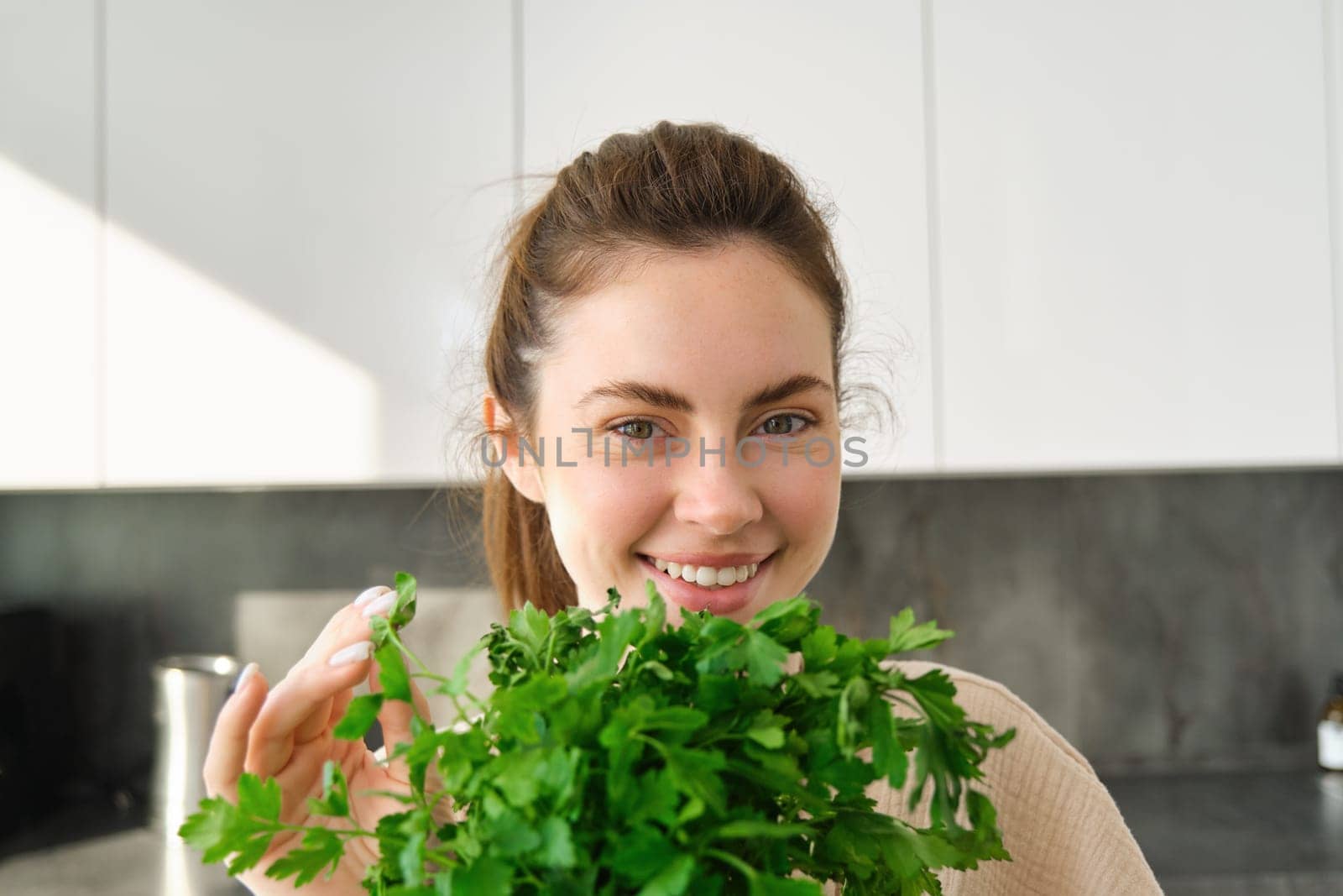 Portrait of beautiful smiling girl with bouquet of parsley, standing in the kitchen and cooking, adding herbs to healthy fresh salad or meal, preparing food by Benzoix