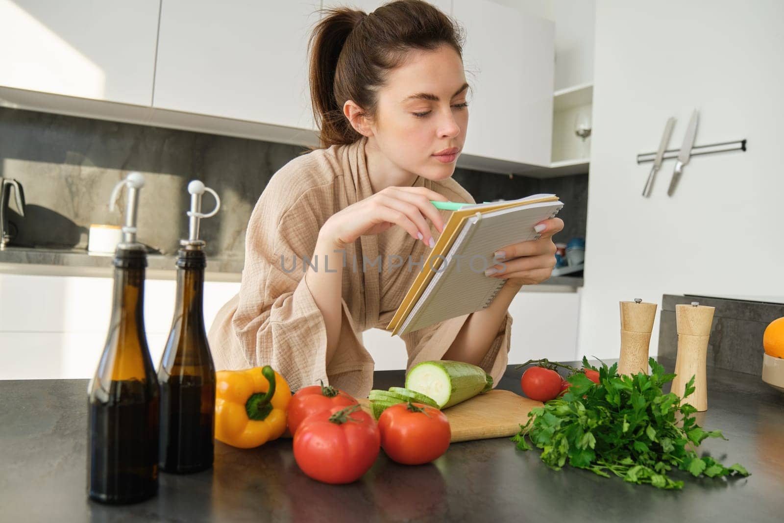Portrait of woman checking grocery list, looking at vegetables, holding notebook, reading recipe while cooking meal in the kitchen, chopping tomatoes and zucchini by Benzoix