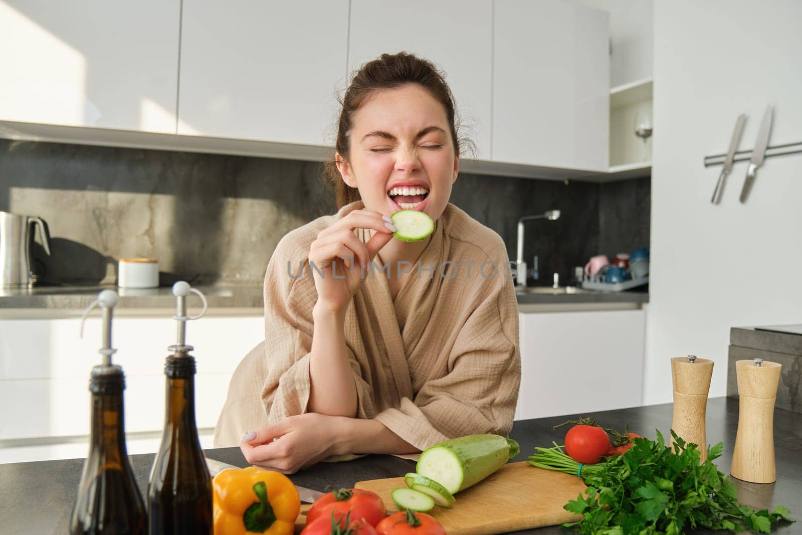 Portrait of happy girl eating vegetables while making meal, cooking healthy vegetarian food in kitchen, holding zucchini, posing in bathrobe by Benzoix