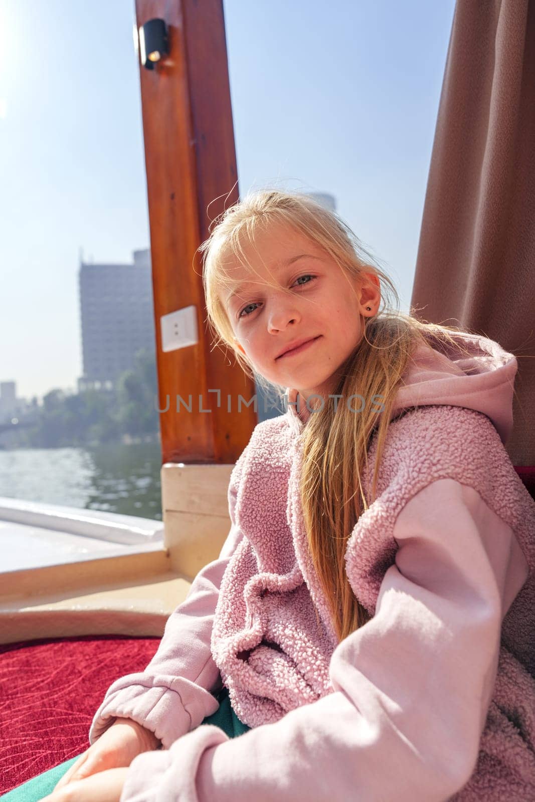 A girl relaxes on a boat, dressed in a cozy pink outfit, looking at the camera. The sunny city skyline and calm waters create a serene atmosphere around her.