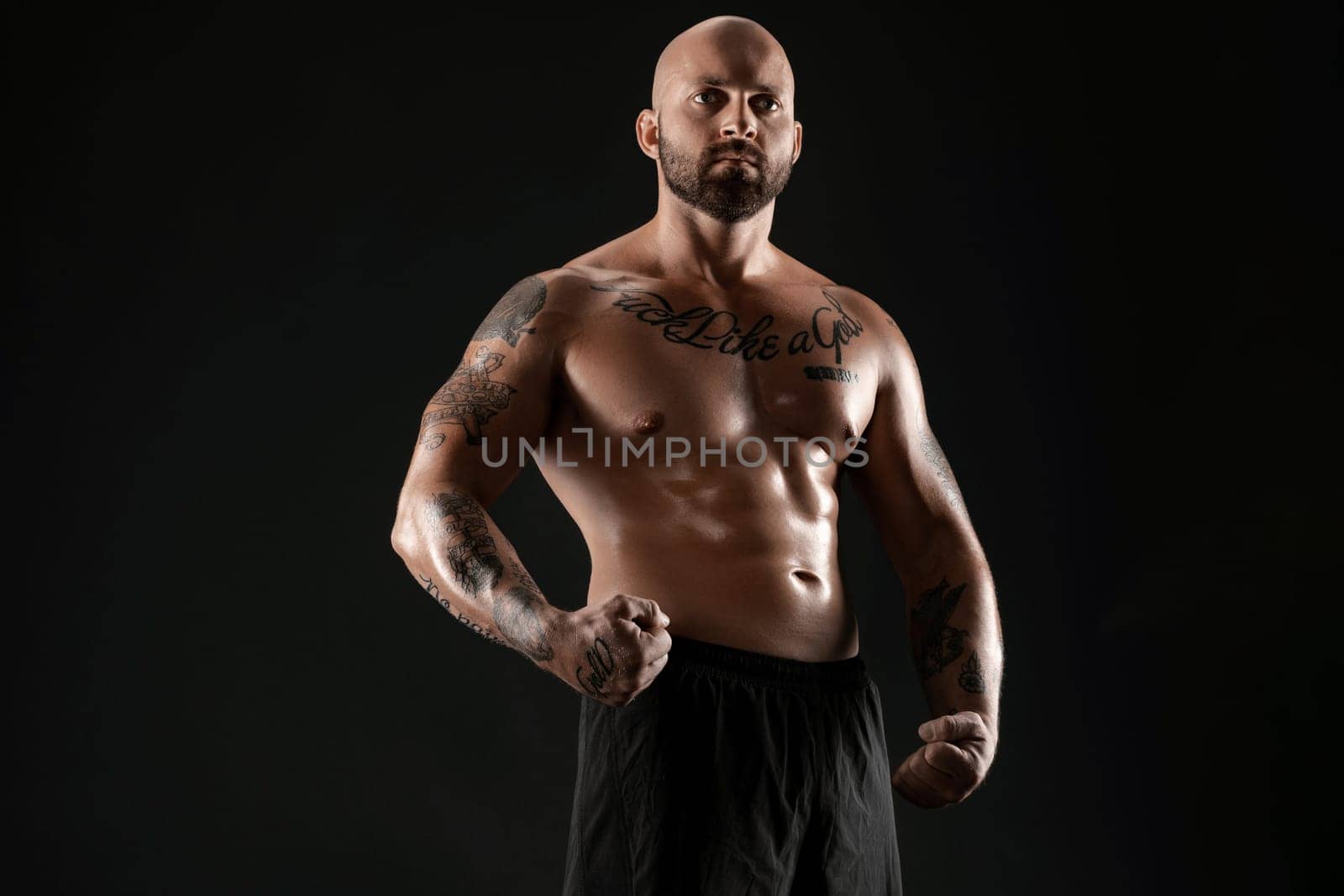 Handsome bald, bearded, tattooed fellow in black shorts is demonstating his muscles posing against a black background and looking at the camera. Chic muscular body, fitness, gym, healthy lifestyle concept. Close-up portrait.