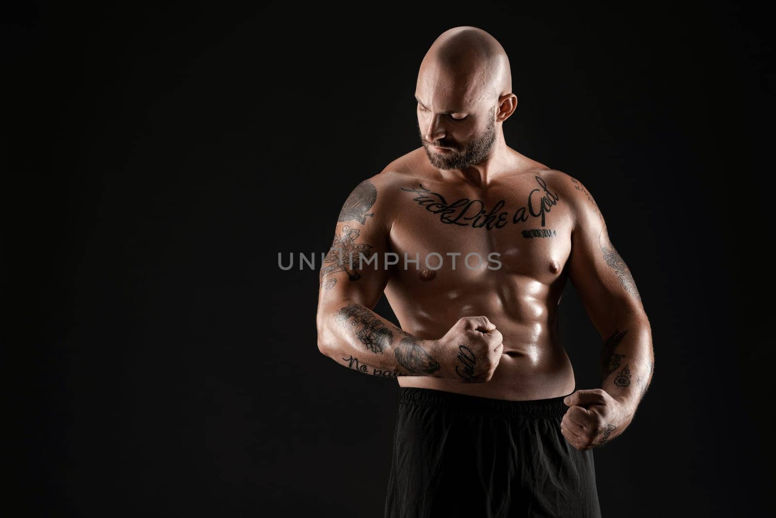 Handsome bald, bearded, tattooed person in black shorts is demonstating his muscles and looking at it while posing against a black background. Chic muscular body, fitness, gym, healthy lifestyle concept. Close-up portrait.