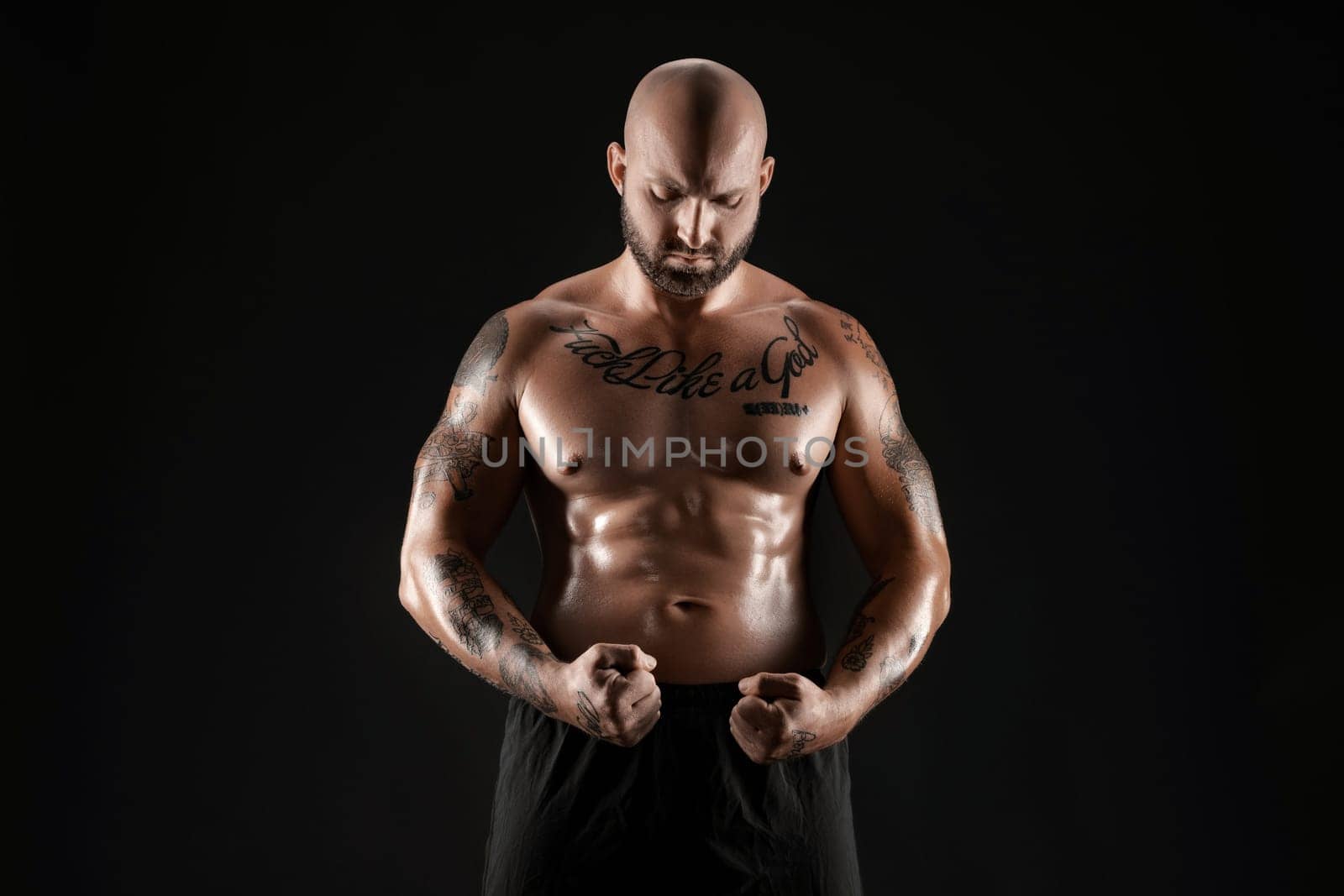 Good-looking bald, bearded, tattooed man in black shorts is demonstating his muscles posing against a black background and looking down. Chic muscular body, fitness, gym, healthy lifestyle concept. Close-up portrait.