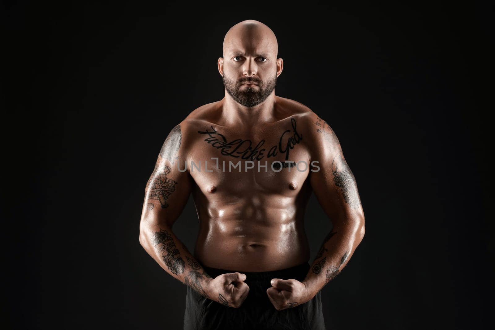 Good-looking bald, bearded, tattooed male in black shorts is demonstating his muscles posing against a black background and looking at the camera. Chic muscular body, fitness, gym, healthy lifestyle concept. Close-up portrait.