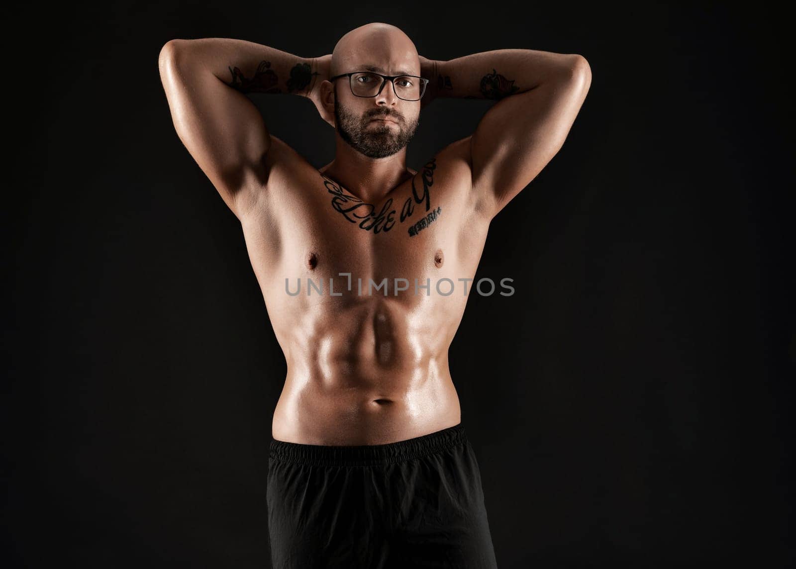 Good-looking bald, bearded, tattooed guy in black shorts is demonstating his muscles while posing against a black background and looking at the camera. Chic muscular body, fitness, gym, healthy lifestyle concept. Close-up portrait.