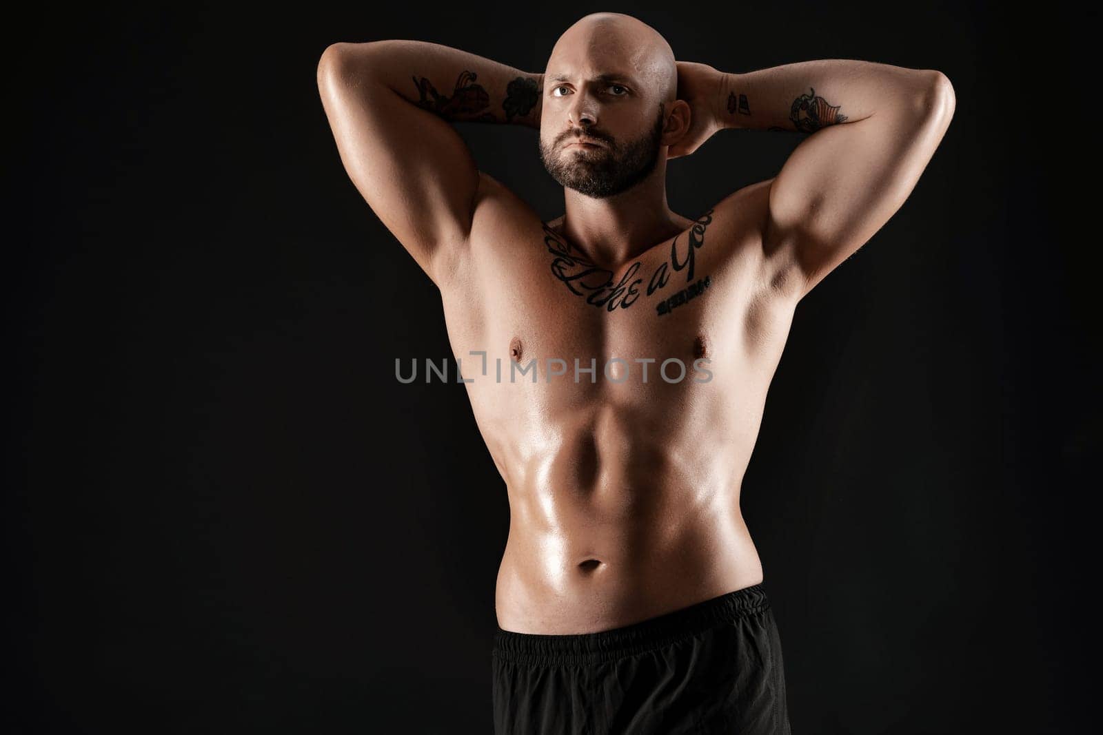 Good-looking bald, bearded, tattooed fellow in black shorts is demonstating his muscles while posing against a black background and looking away. Chic muscular body, fitness, gym, healthy lifestyle concept. Close-up portrait.