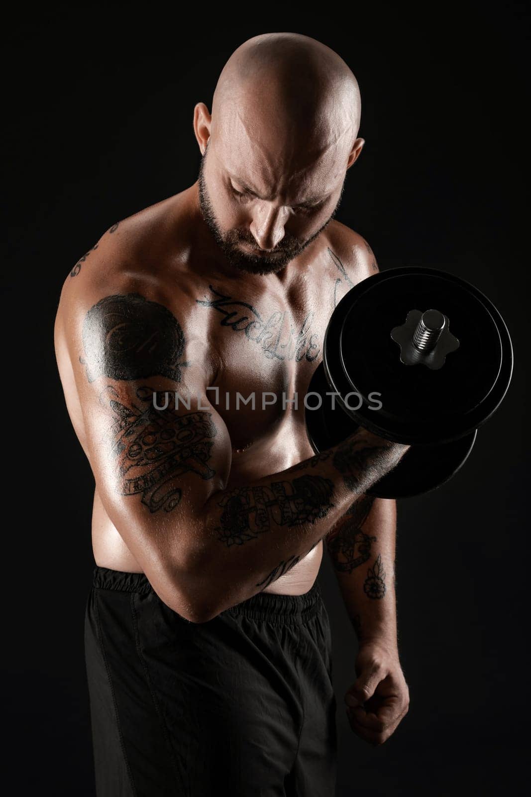 Athletic bald, bearded, tattooed man in black shorts is posing with a dumbbell against a black background. Close-up portrait. by nazarovsergey