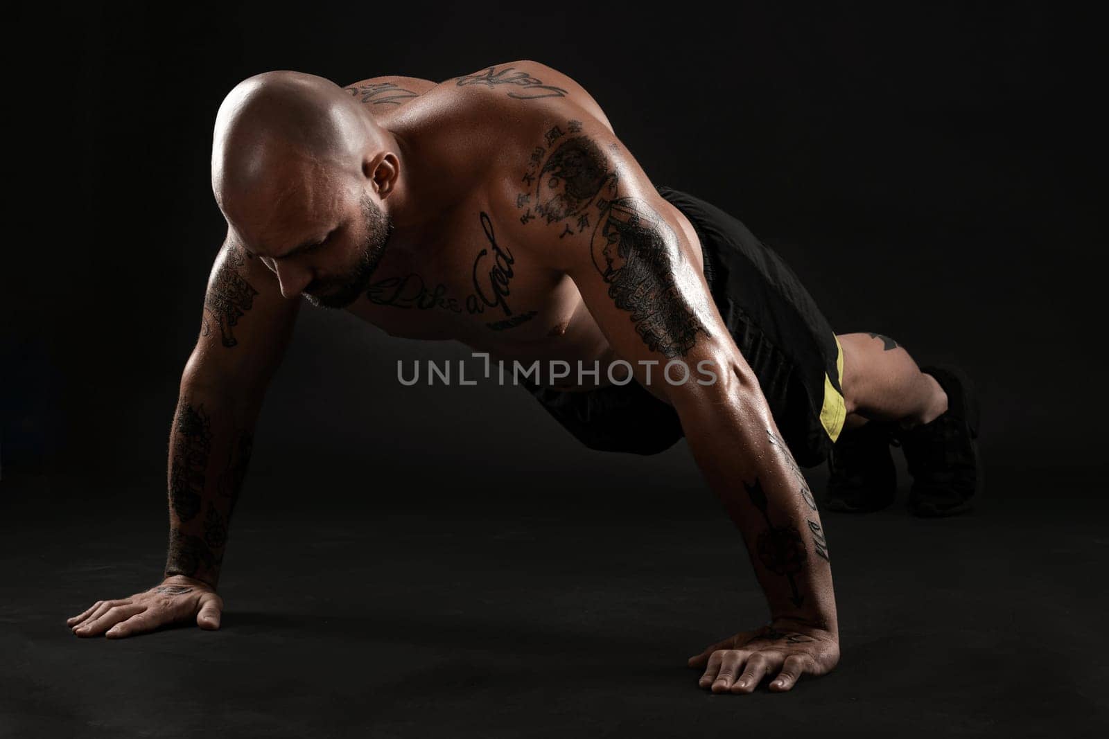 Stately bald, bearded, tattooed man in black shorts and sneakers is pushing ups from the floor against a black background. Chic muscular body, fitness, gym, healthy lifestyle concept. Close-up portrait.