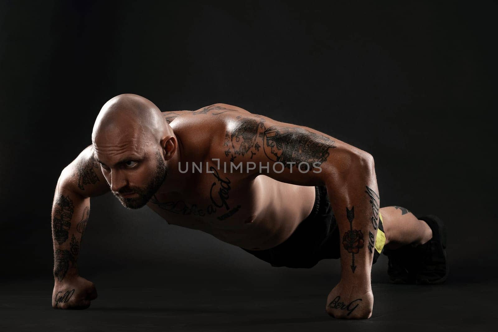 Stately bald, bearded, tattooed male in black shorts and sneakers is pushing ups from the floor against a black background. Chic muscular body, fitness, gym, healthy lifestyle concept. Close-up portrait.