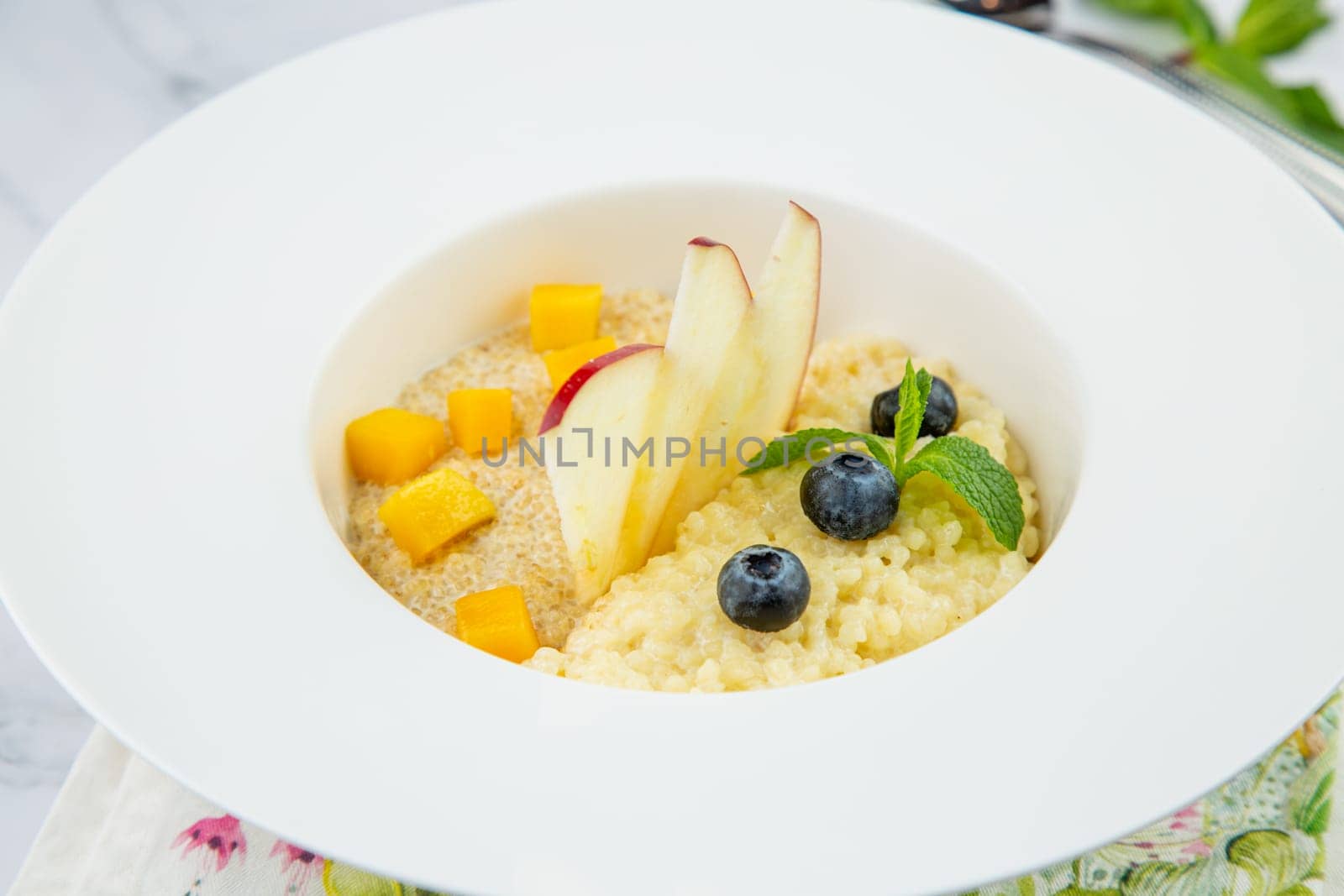 porridge with pieces of apple, mango, berries and mint in a white plate side view by tewolf