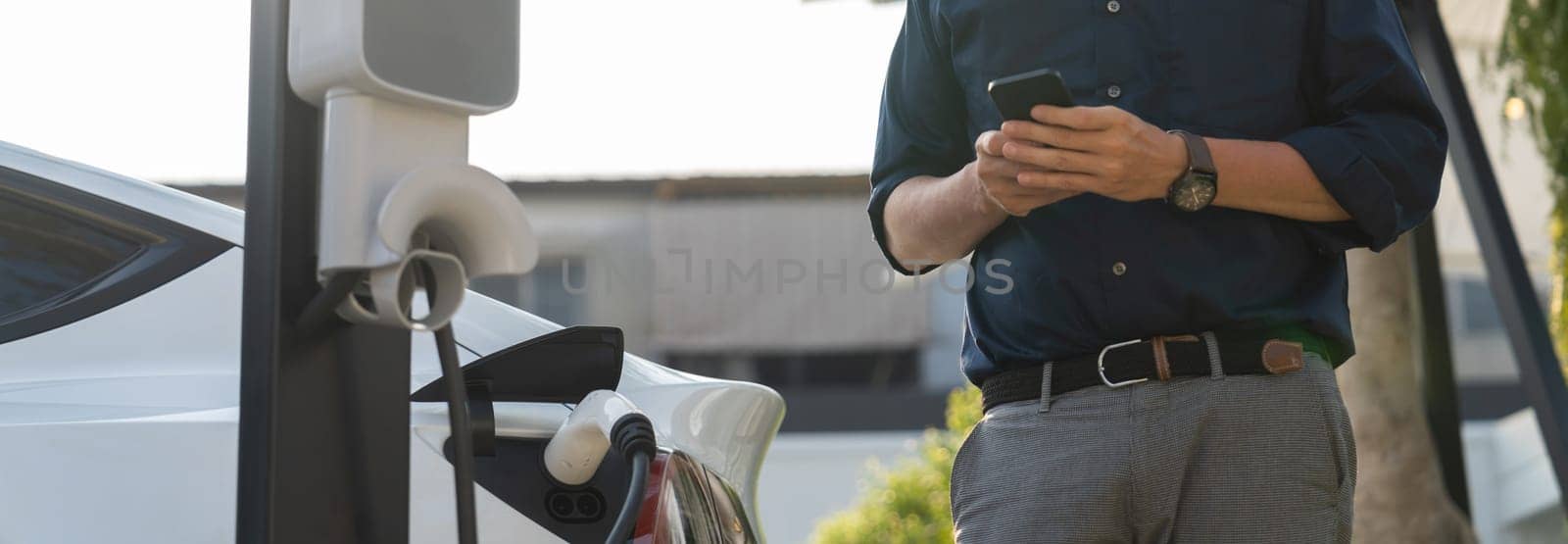 Young man use smartphone to pay for electricity at public EV car charging station at city commercial mall parking lot. Modern environmental and sustainable urban lifestyle with EV vehicle. Expedient