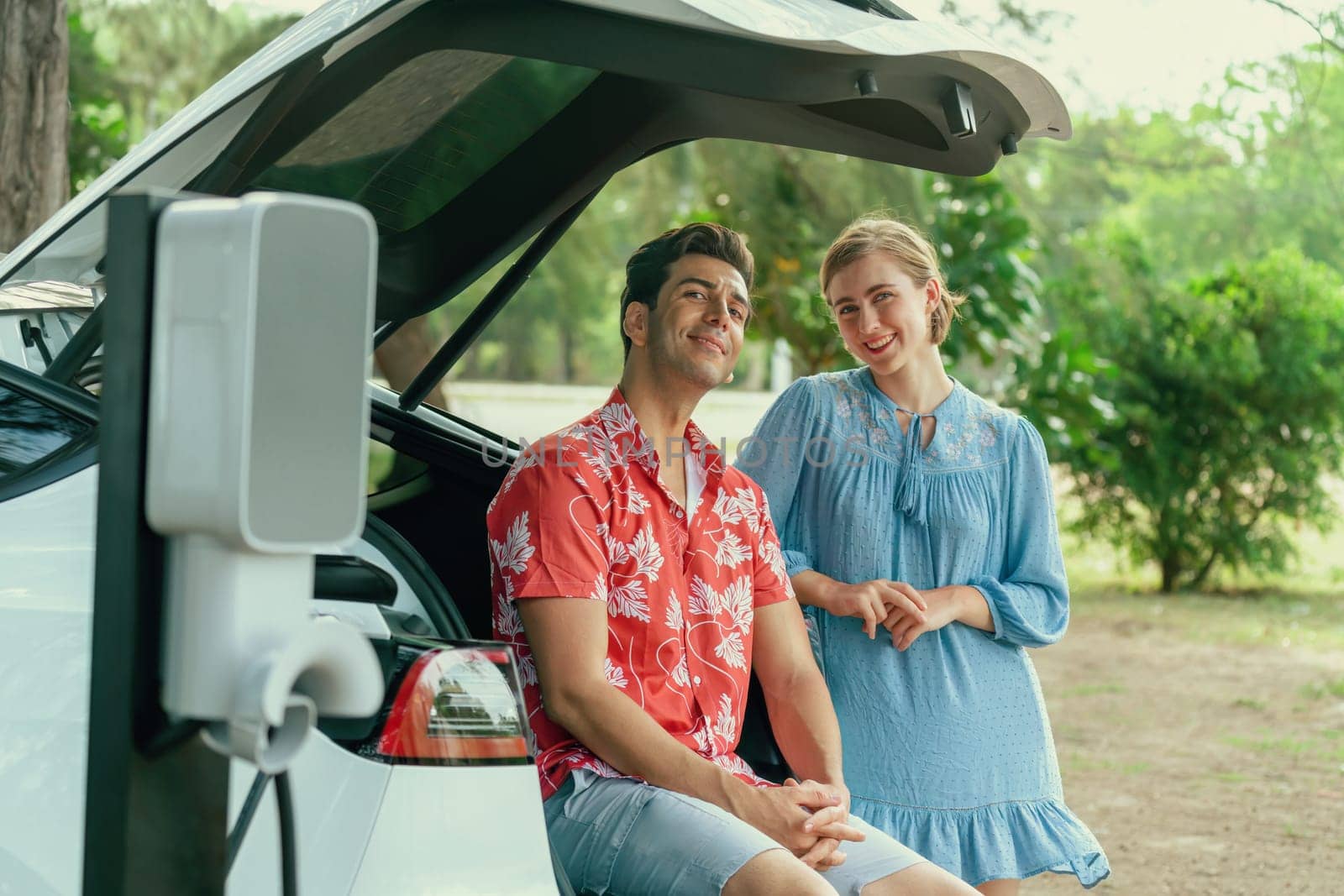 Road trip vacation with electric vehicle, lovely couple sitting on trunk and recharge EV car with green and clean energy. Nature and travel with eco-friendly car for sustainable environment. Perpetual