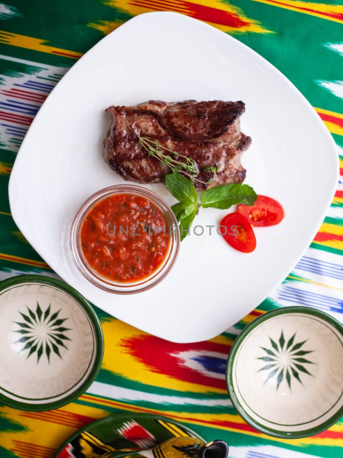 beef steak with tomato sauce, tomatoes and herbs in oriental style on a table with a teapot and a cup for tea. by tewolf