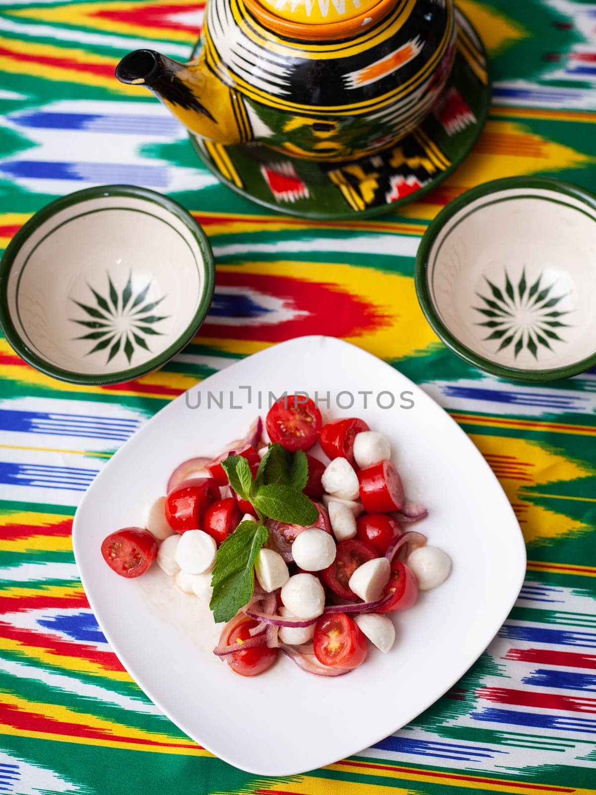 caprese salad with mazzarella tomatoes and herbs in oriental style on a table with a teapot and a cup for tea. by tewolf