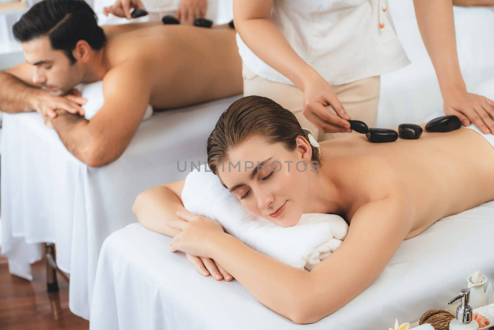 Hot stone massage at spa salon in luxury resort with day light serenity ambient, blissful couple customer enjoying spa basalt stone massage glide over body with soothing warmth. Quiescent