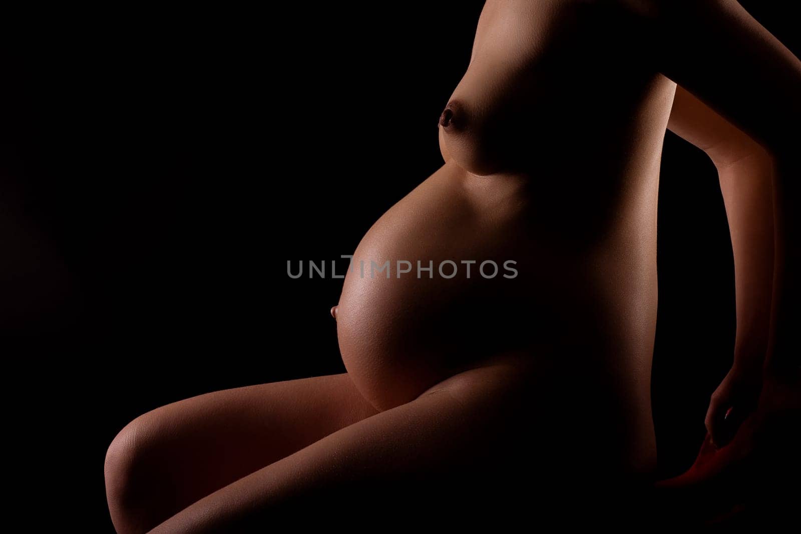 Pregnant attractive woman (8th month) leaning on sitting. Shoulders to legs visible. Artistically illuminated. Black background.