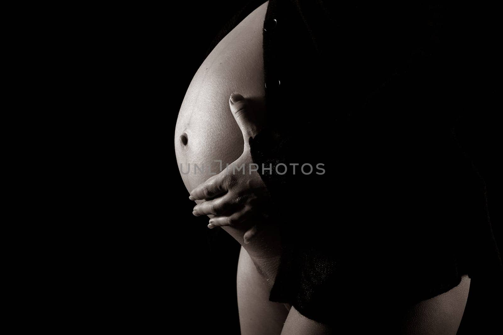 Pregnant attractive woman (8th month) holding her stomach. Black jacket. Monochrome. Black background.