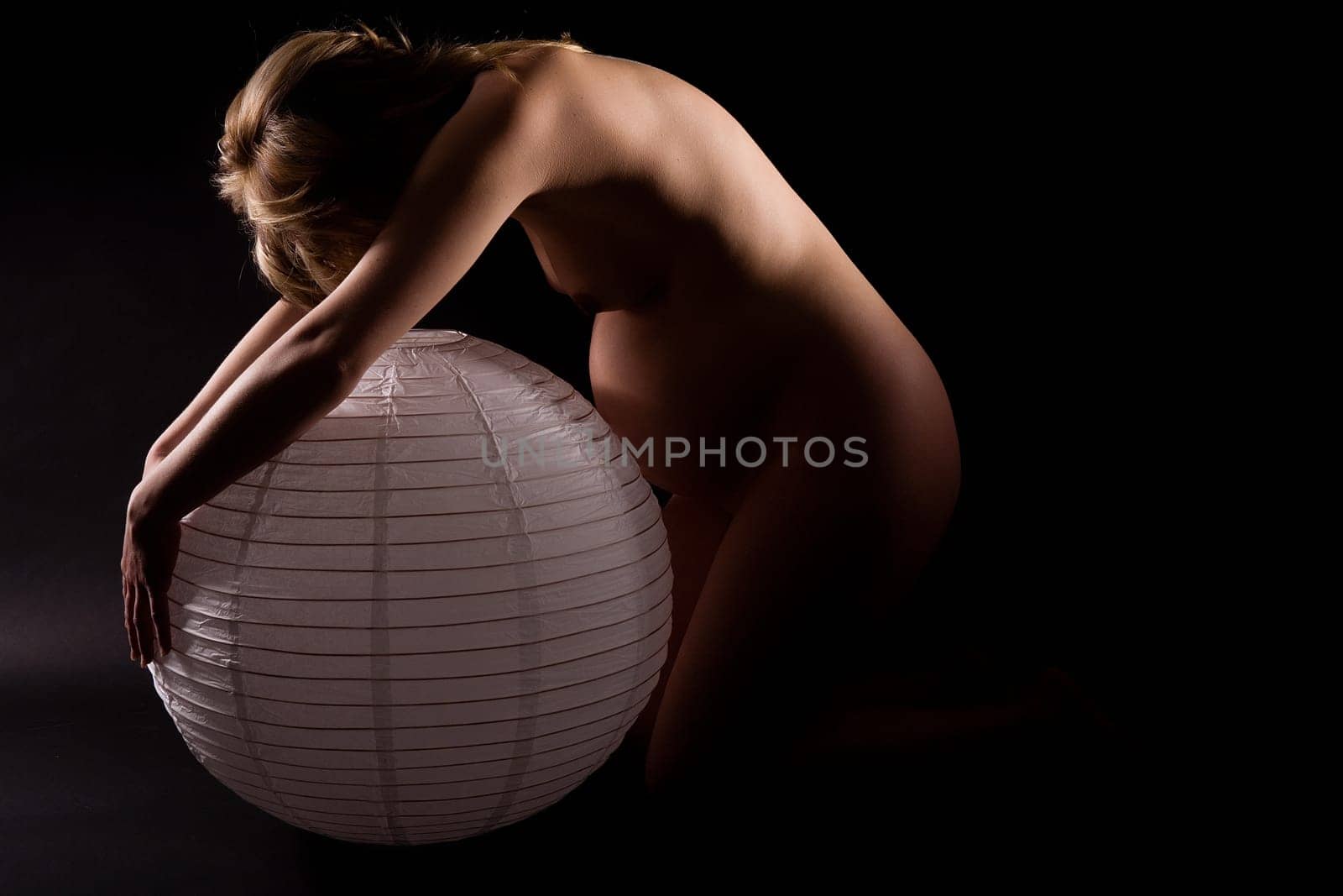 Pregnant attractive woman (8th month) artistically bending over paper lamp. Belly comparison... Black background.