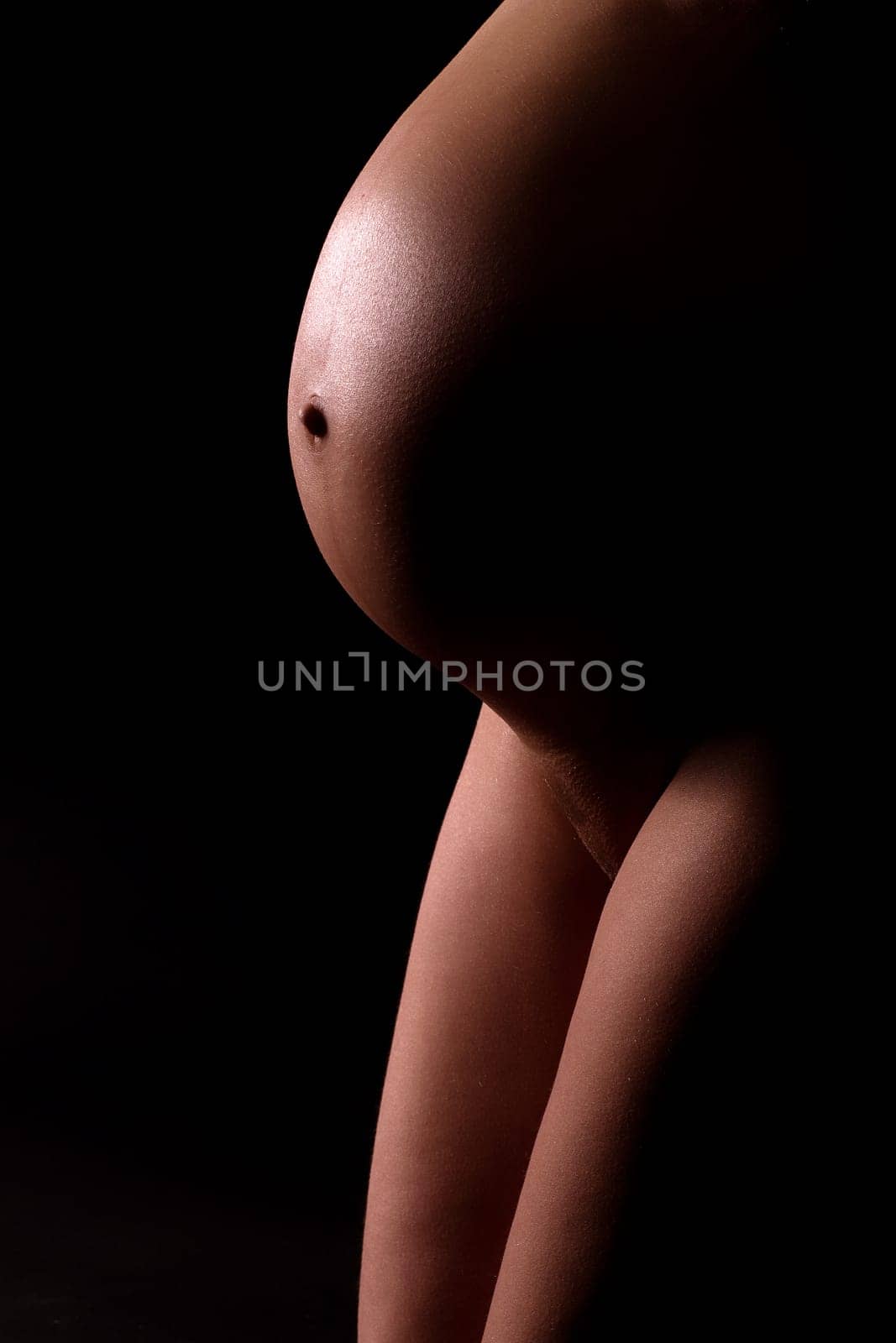 Baby bump and thighs. Pregnant attractive woman (8th month). Artistically illuminated. Black background.