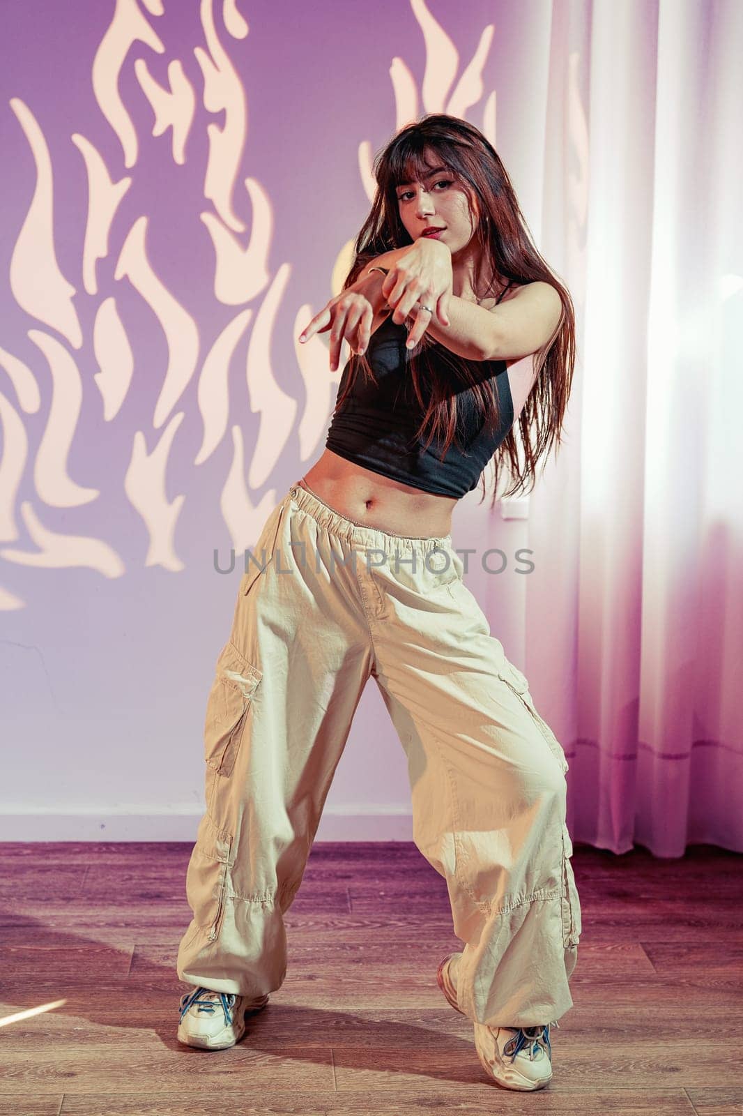 A dancer performs a vibrant routine in a contemporary studio, wearing light-colored baggy pants and a fitted top. The backdrop features abstract flames, enhancing the energetic atmosphere.