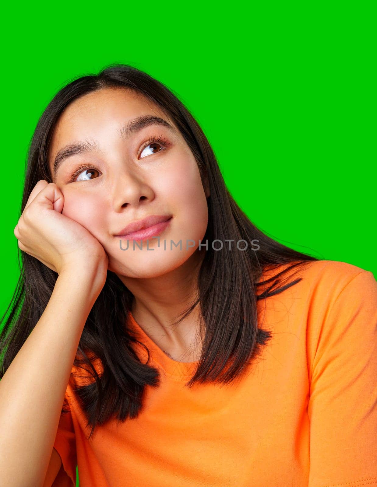 A young woman is seen wearing a vibrant orange shirt, resting her chin on her hand while looking thoughtfully to the side. The bright green background enhances her casual pose.