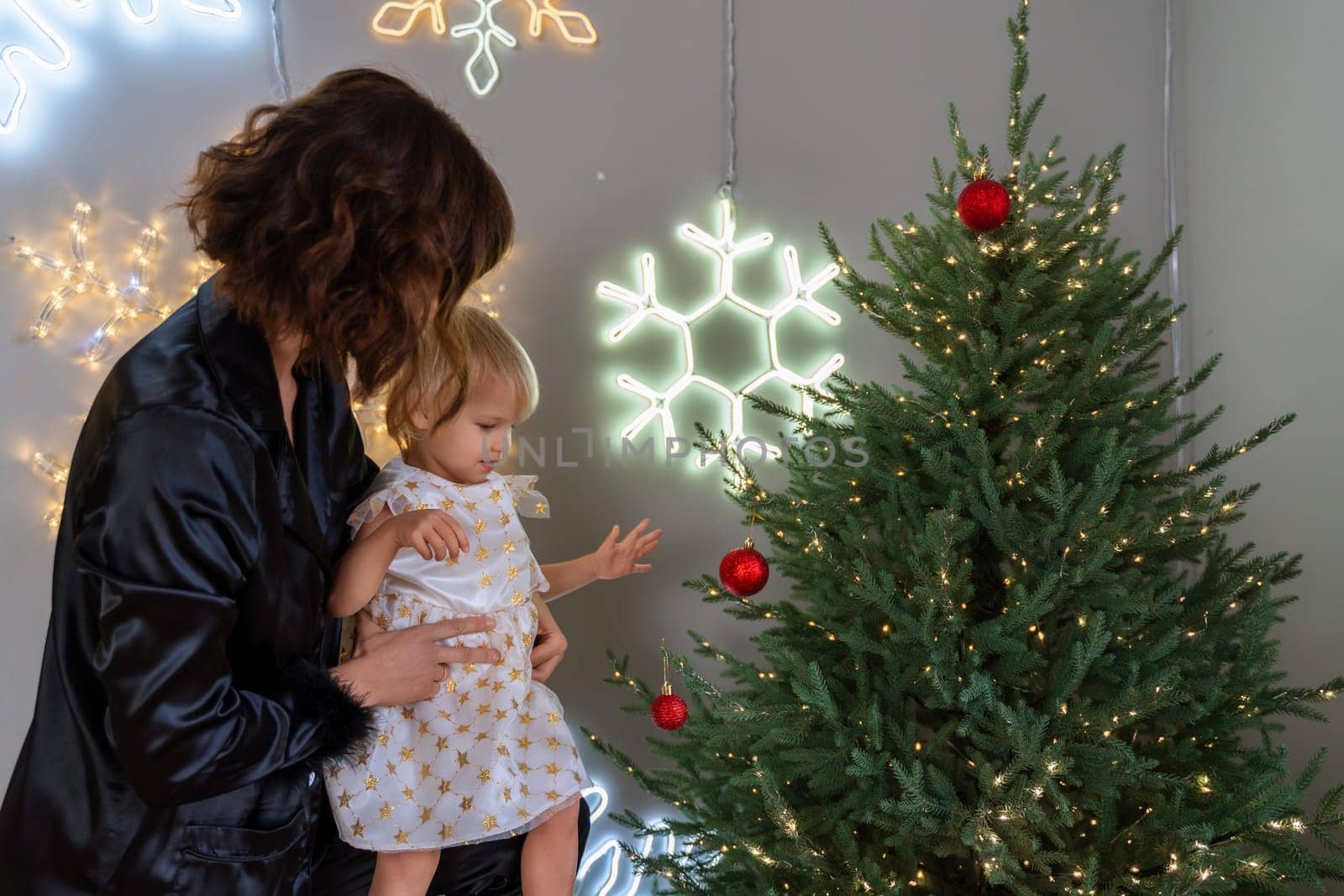 A mother with a 2-year-old daughter decorates the Christmas tree. Mom in a black suit, a girl in a white dress, her daughter hangs a red ball on the Christmas tree. Merry Christmas and New Year concept by Matiunina