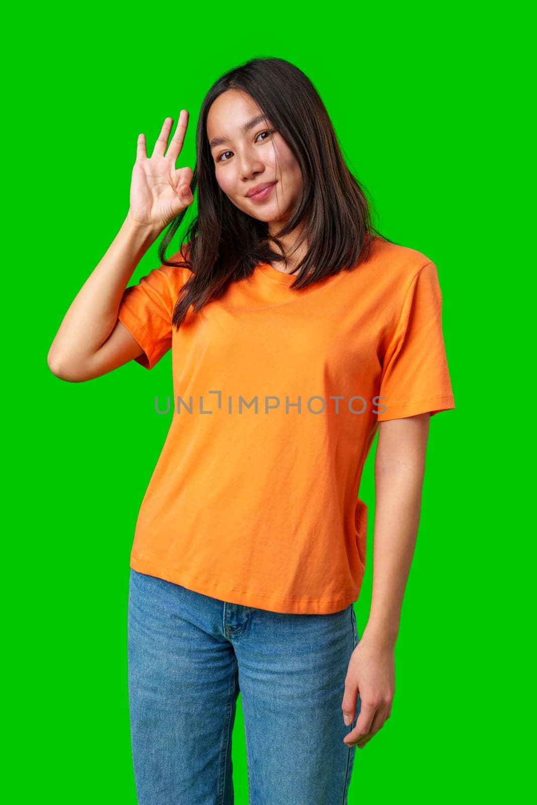 A young woman stands confidently with a smile, wearing a bright orange shirt and blue jeans. Her hand is raised in a playful gesture, set against a vibrant green backdrop.