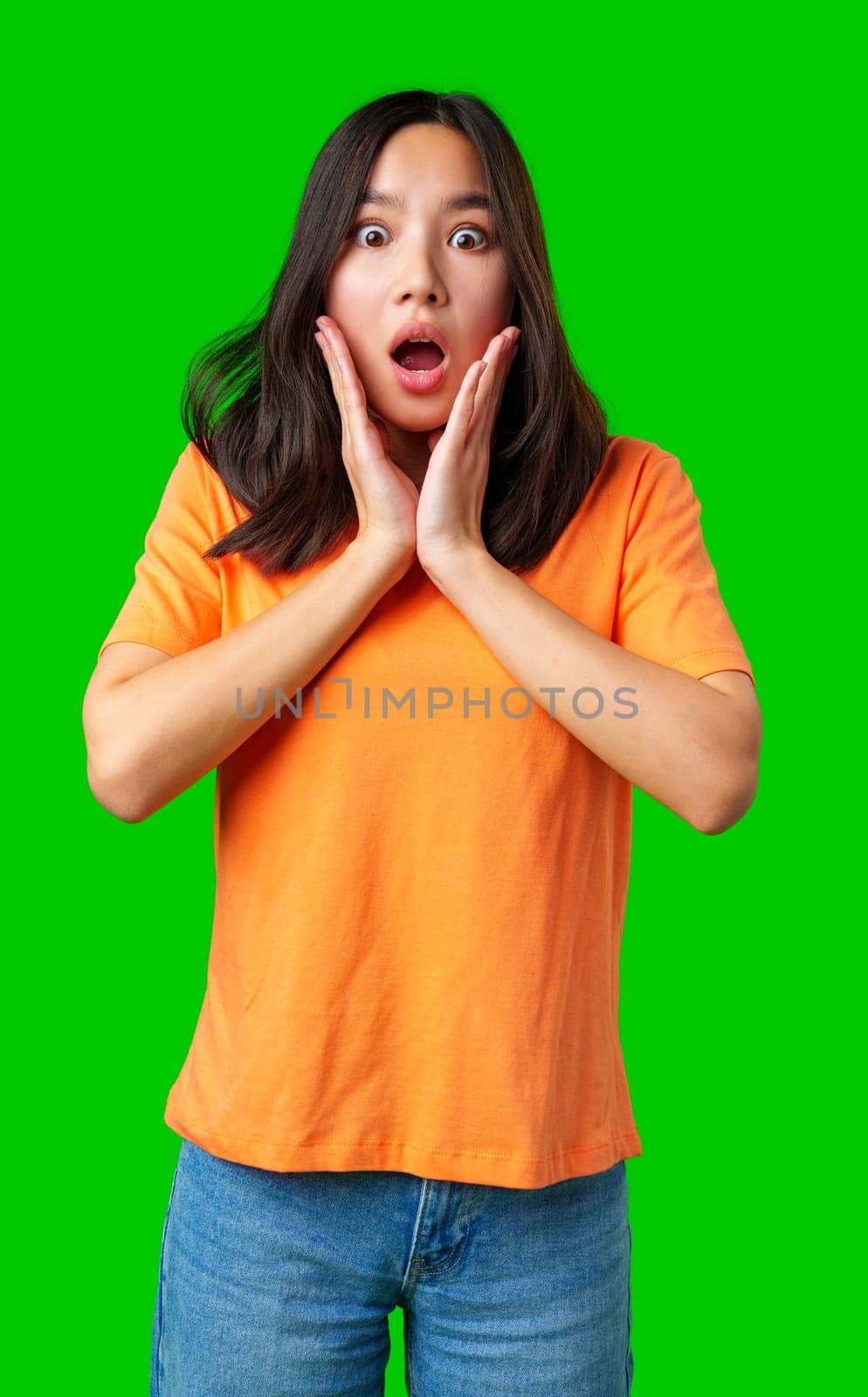 A young woman with long dark hair is wearing an orange shirt and denim jeans. She shows a surprised expression while holding her face, highlighting her astonishment.