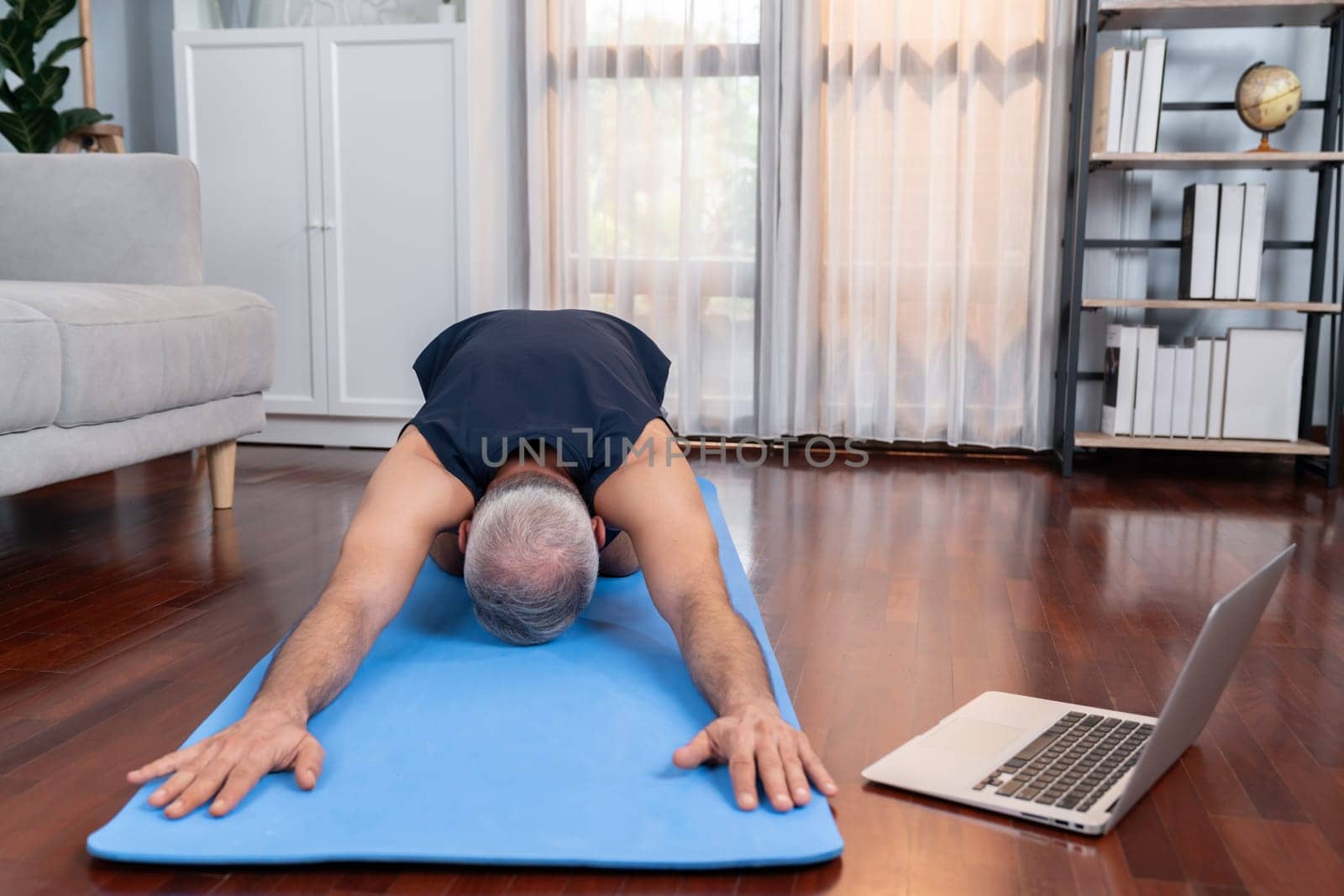 Senior man in sportswear being doing yoga in meditation posture on exercising mat at home. Healthy senior pensioner lifestyle with peaceful mind and serenity. Clout