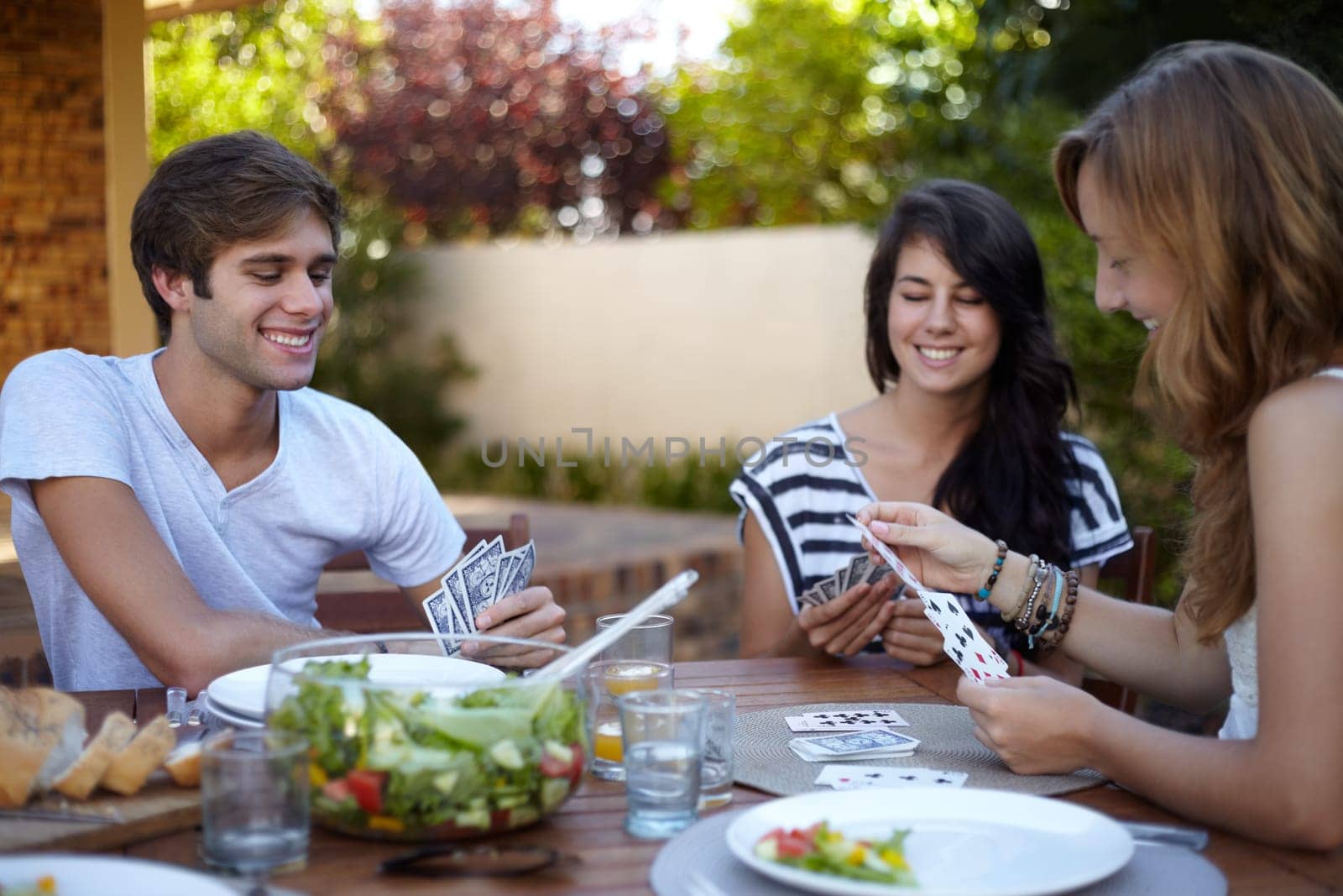 Friends, card game and food in backyard for lunch and discussion with a smile and fun. Salad, laughing and young people outdoor of a home with communication and funny joke together with a meal by YuriArcurs