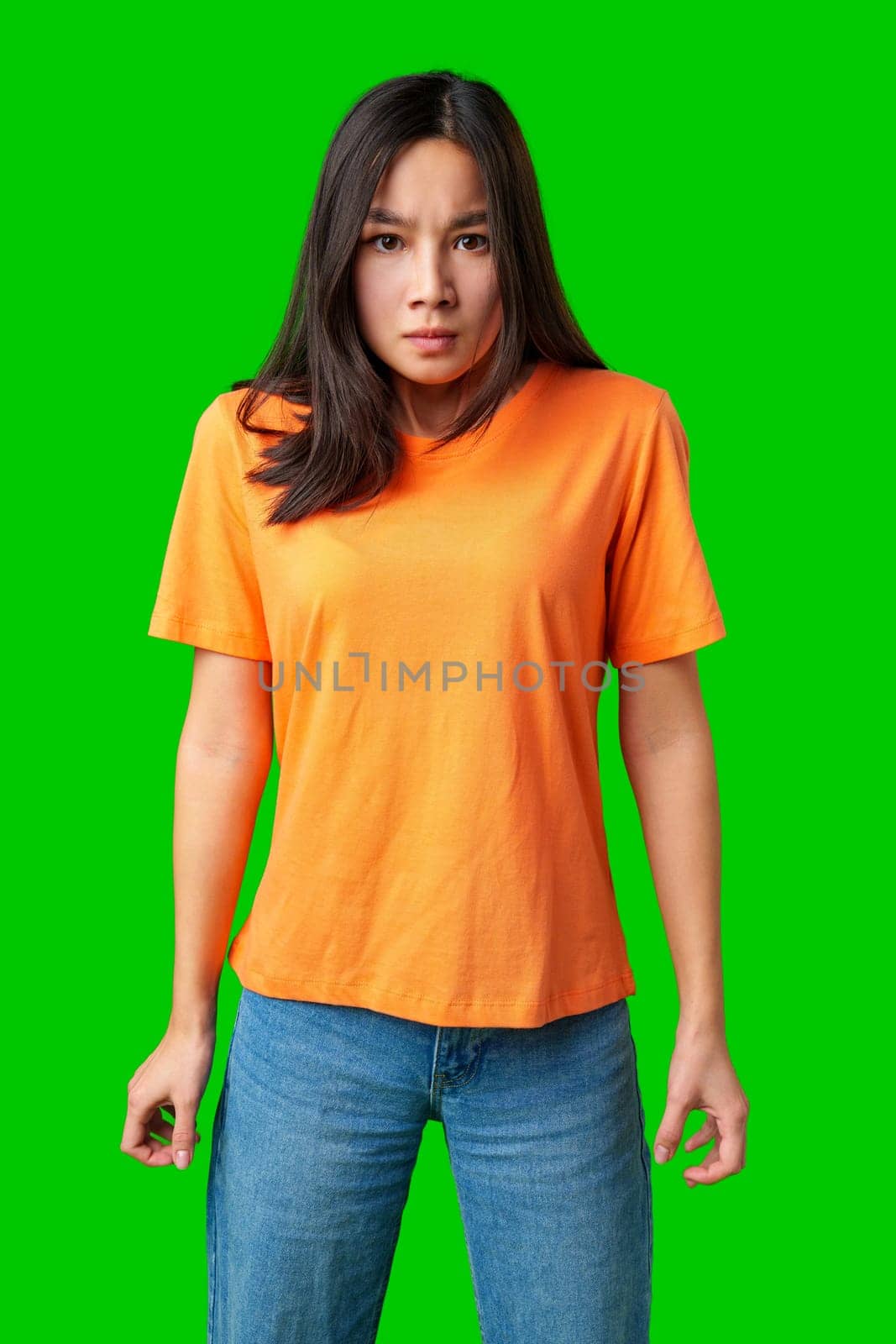 A woman stands confidently with her hands clenched, wearing a bright orange shirt and blue jeans. Her expression reflects determination and strength against a vivid green backdrop.