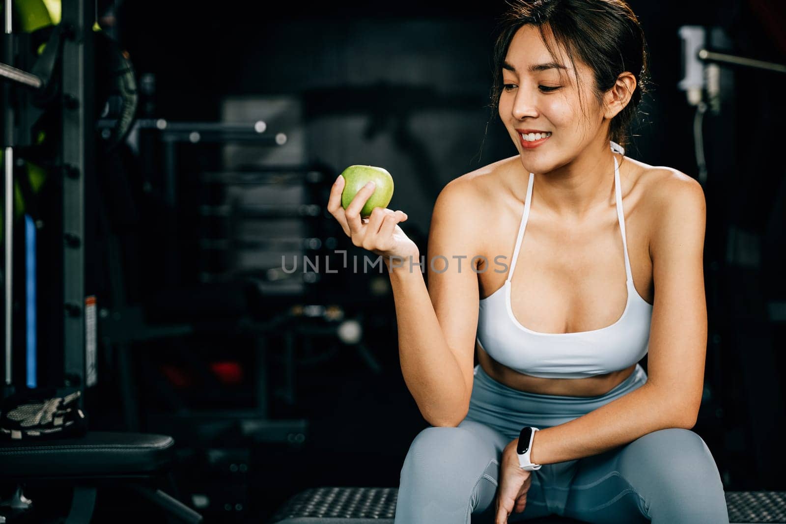 An Asian girl holds a green apple in a fitness gym, emphasizing the importance of healthy food choices for maintaining a slim and sportive physique. Healthy fitness and eating lifestyle concept