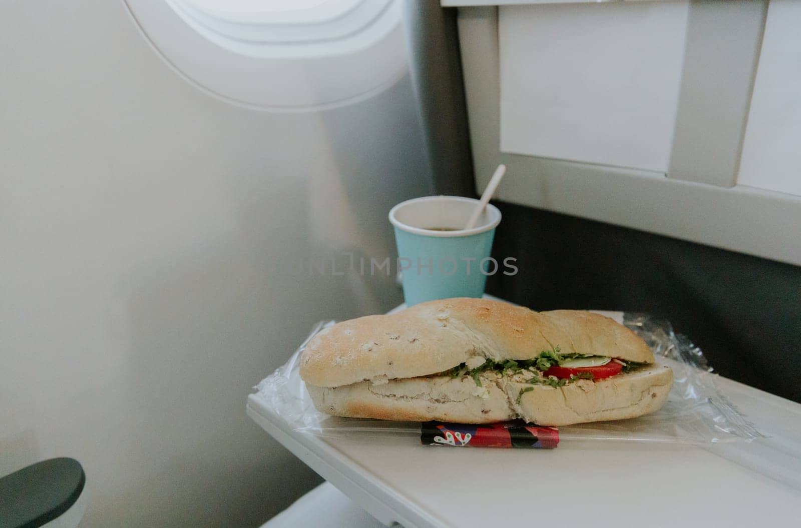 One appetizing sandwich with tuna, tomatoes and a paper glass of tea lie on a table on the plane, close-up side view.