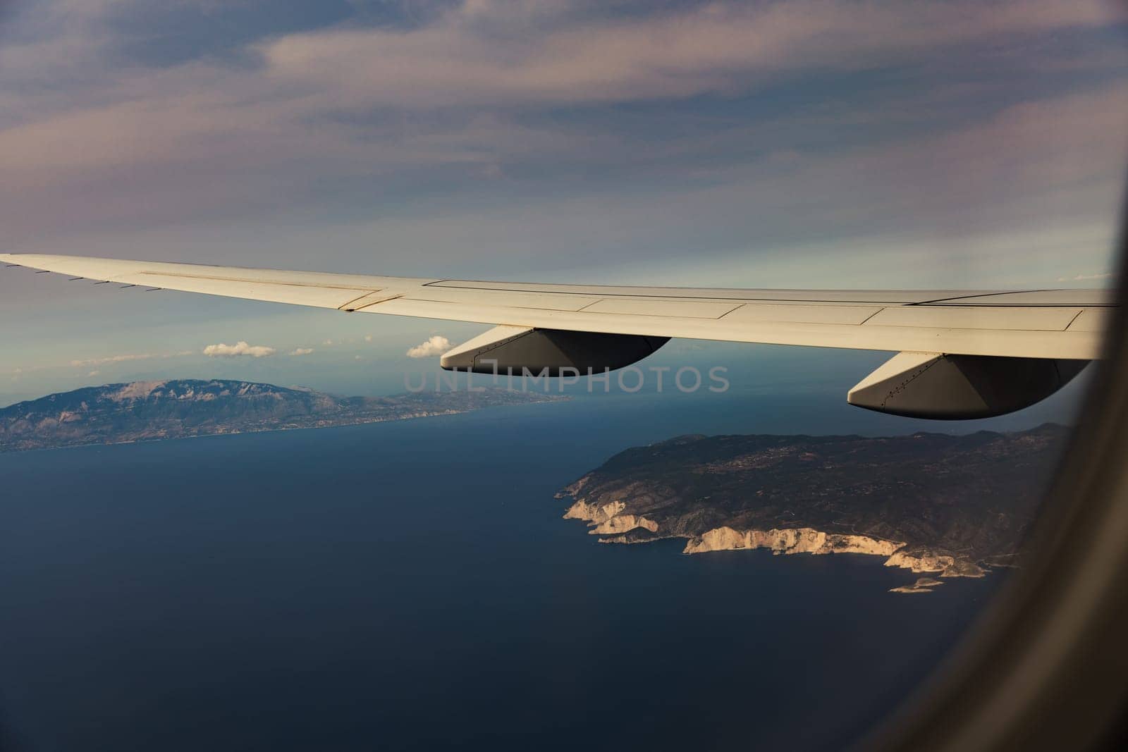 A beautiful view of the white wing of an airplane against the backdrop of a blurred island in the blue sea through the window in the evening summer during a flight, close-up side view.