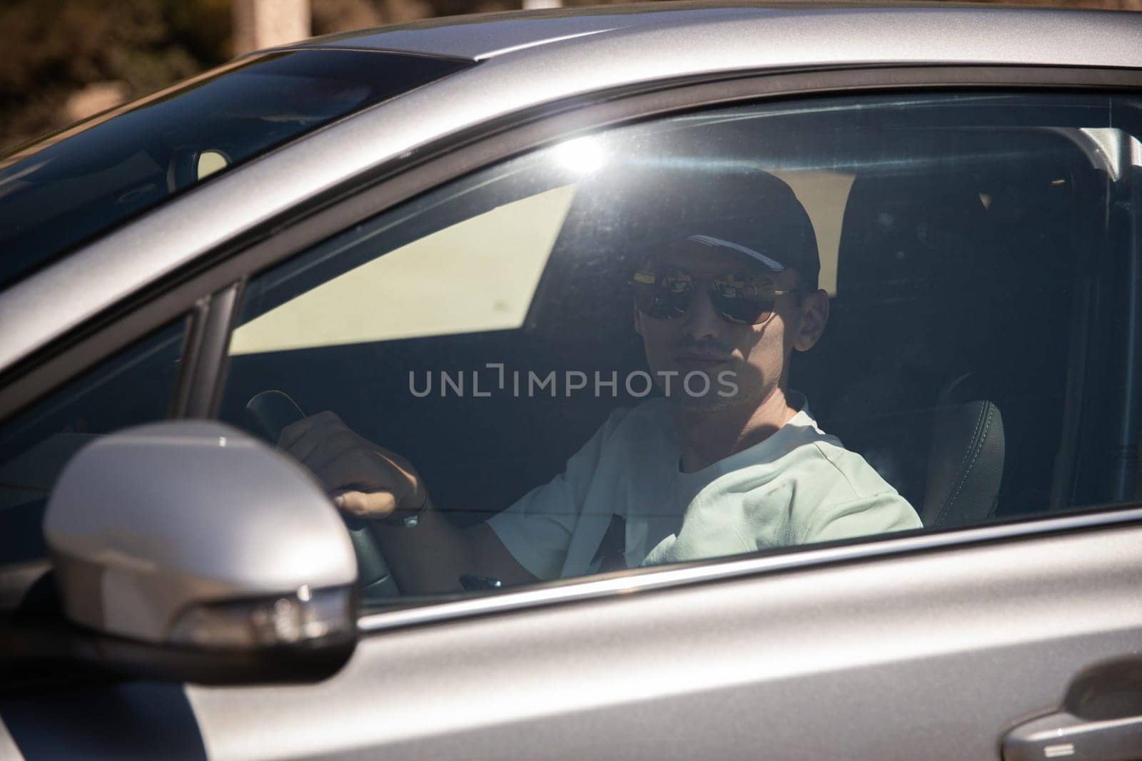 One young handsome Caucasian guy in a cap and sunglasses sits in a gray car behind the wheel on a sunny summer day, close-up side view.