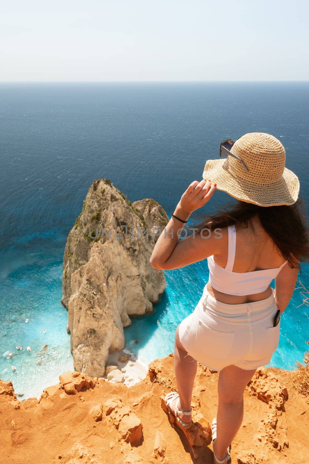 One Caucasian girl in white clothes and a straw hat stands from her back on the edge of the mountain and looks at a rock in the blue sea on a sunny summer day, close-up view from above.