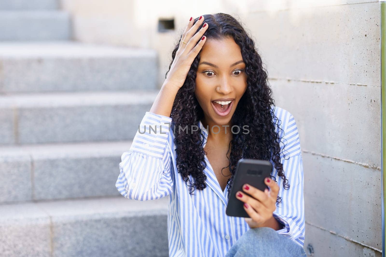 A delighted young woman with beautiful curly hair is sitting on the steps, deeply engaged with her smartphone, filled with surprise and happiness, capturing a unique moment of joy and excitement