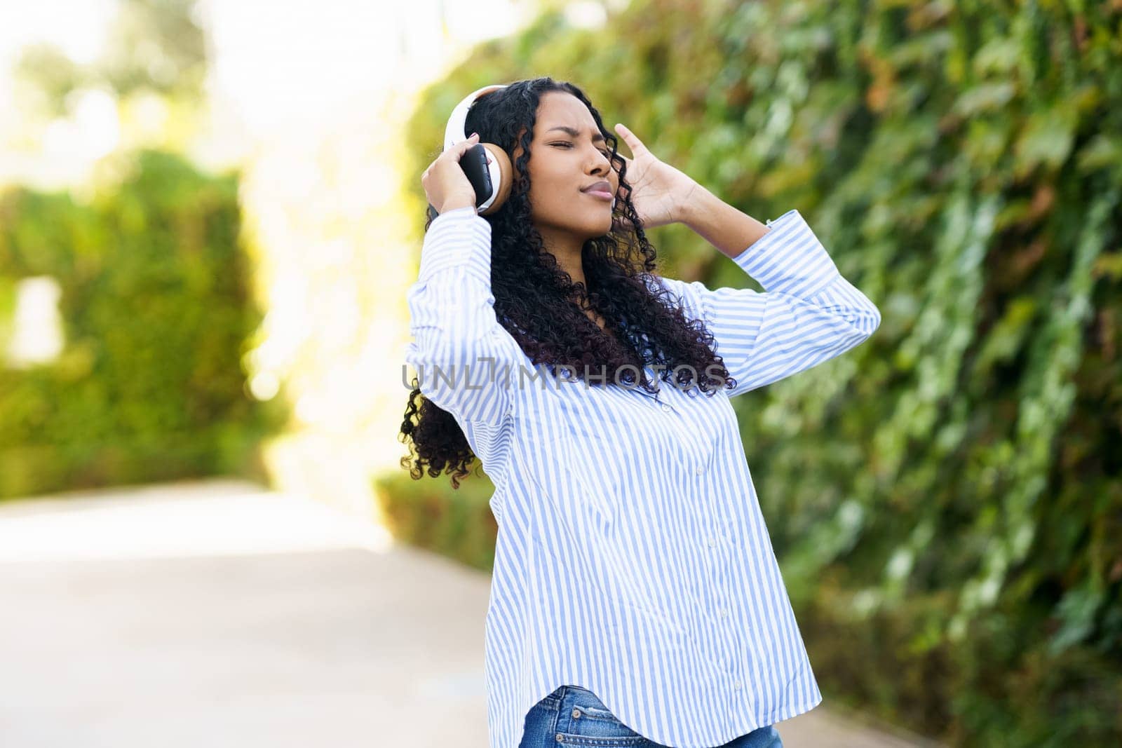 A young woman wearing a stylish striped shirt is enjoying her favorite music through headphones, showing her joy while outdoors, surrounded by vibrant greenery and the beauty of nature