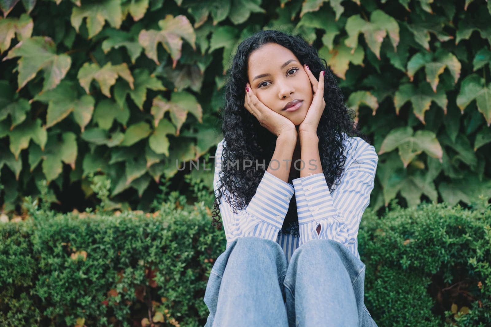 A young woman sits pensively amidst the vibrant and lush green foliage, showcasing her casual yet fashionable style while lost in thought, reflecting her contemplative mood and emotions