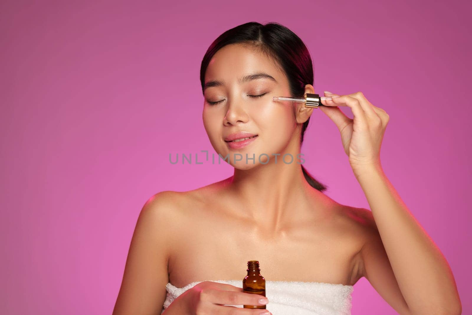 A woman with long hair applies skincare serum to her face while smiling. The vibrant pink background highlights her natural beauty during her daily skincare regimen.