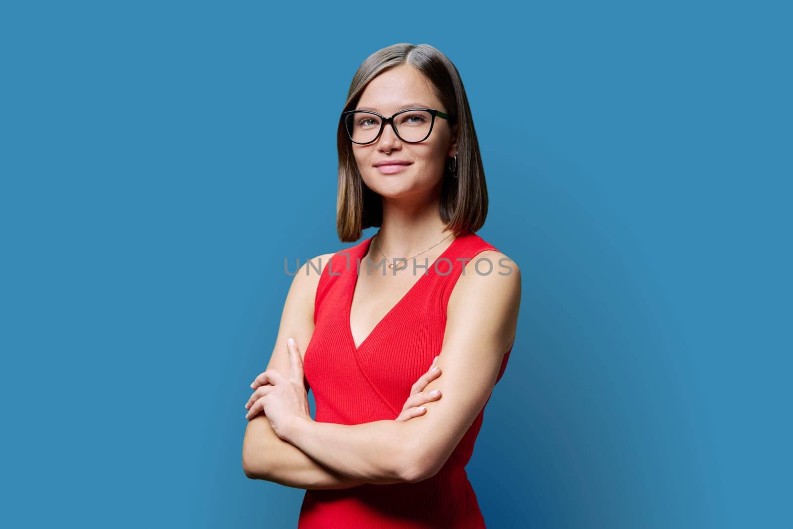 Young confident woman in glasses in red with crossed arms looking at camera on blue studio background. Business, work, services, education, fashion, beauty, professions, people
