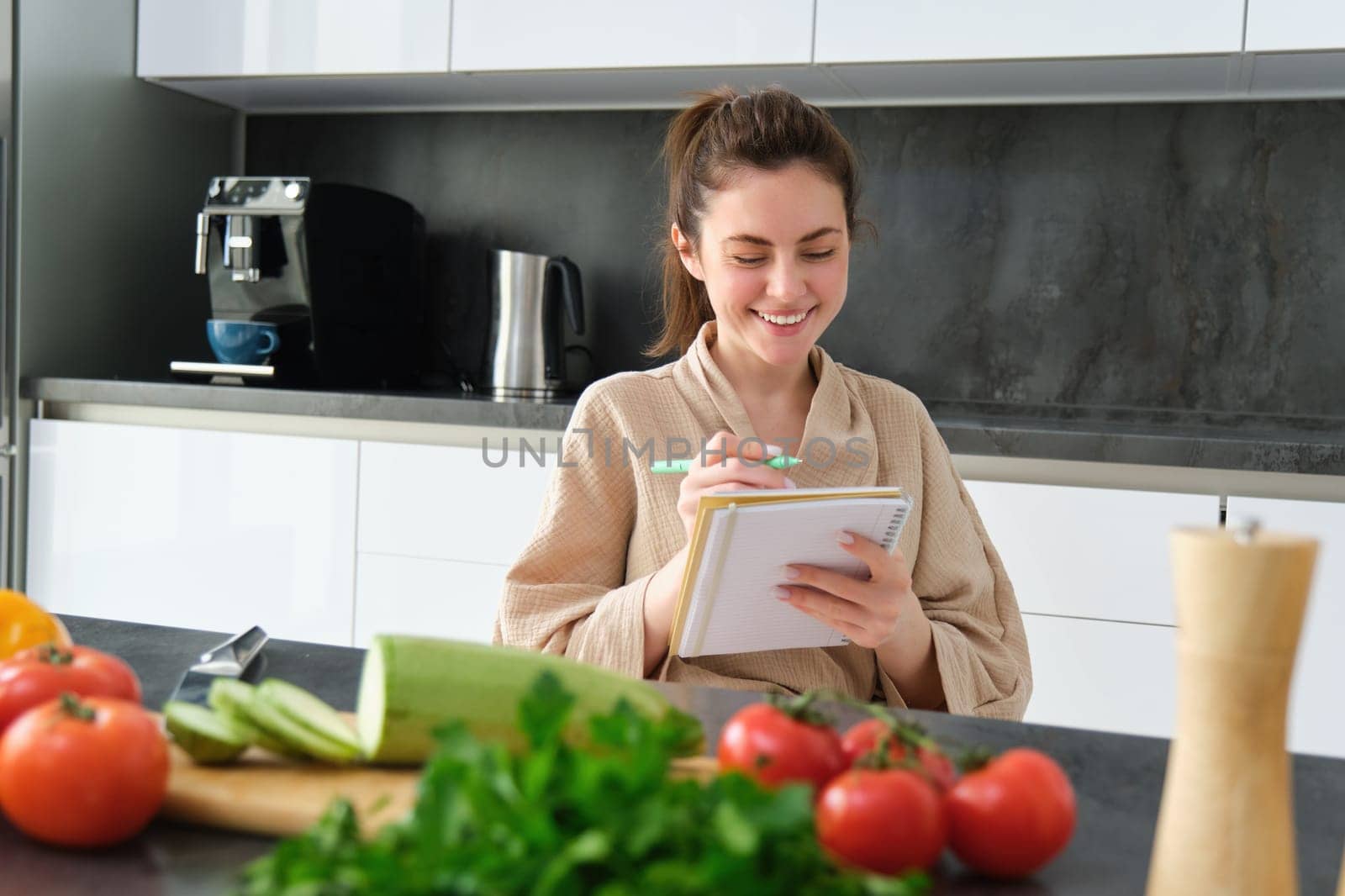 Portrait of beautiful, smiling young woman making list of meals, writing down recipe, sitting in the kitchen with vegetables, doing house errands by Benzoix