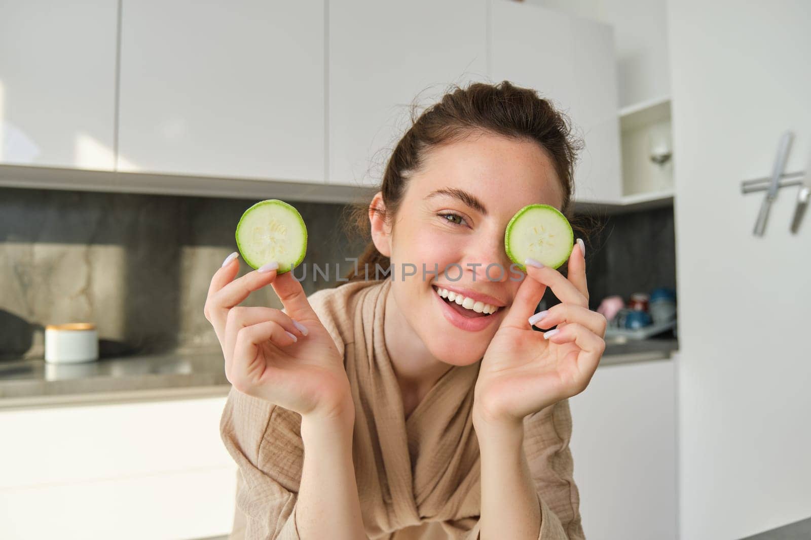 Portrait of beautiful brunette girl cooking in the kitchen, posing in bathrobe at home, holding zucchini, showing happy smile, making healthy food, vegetarian meal.